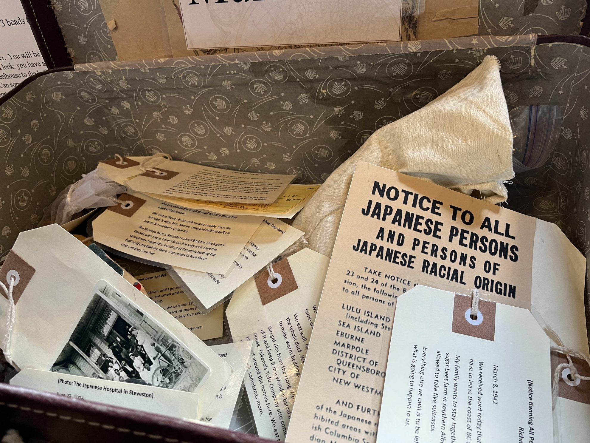 Box made of brown fabric with imperial crown pattern, filled with canvas bas and paper tags, along with a paper with the headline: Notice to all Japanese Persons and Persons of Japanese Racial Origin.