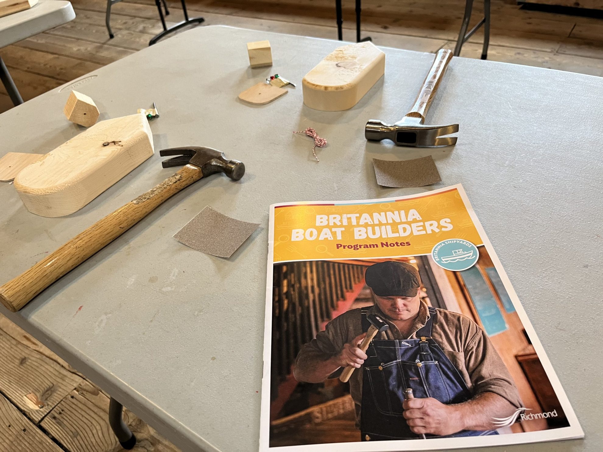 White table with two hammers, blocks of wood and sandpaper, along with a booklet that reads: Britannia Boat Builders Program Notes