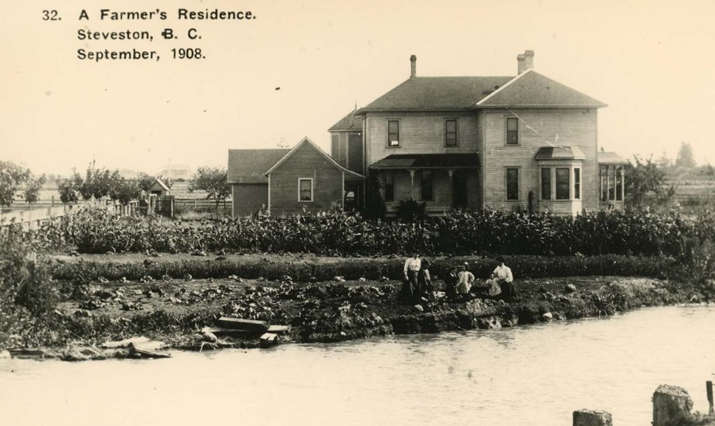 Sepia photo of two wooden buildings standing on water's edge with five people standing on the bank in foreground.