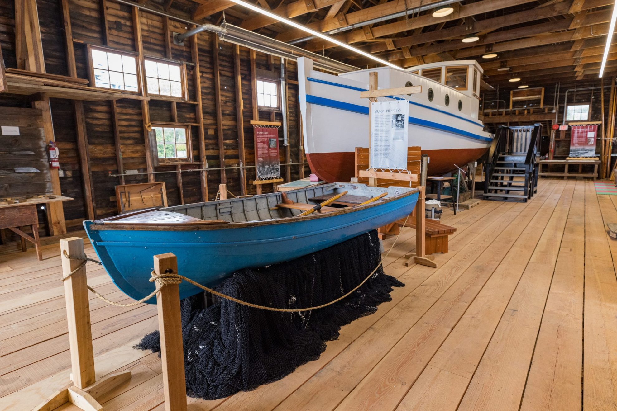 Smaller light blue boat displayed in front of a larger wooden boat that is white with a light blue stripe and red bottom, displayed inside a wooden building