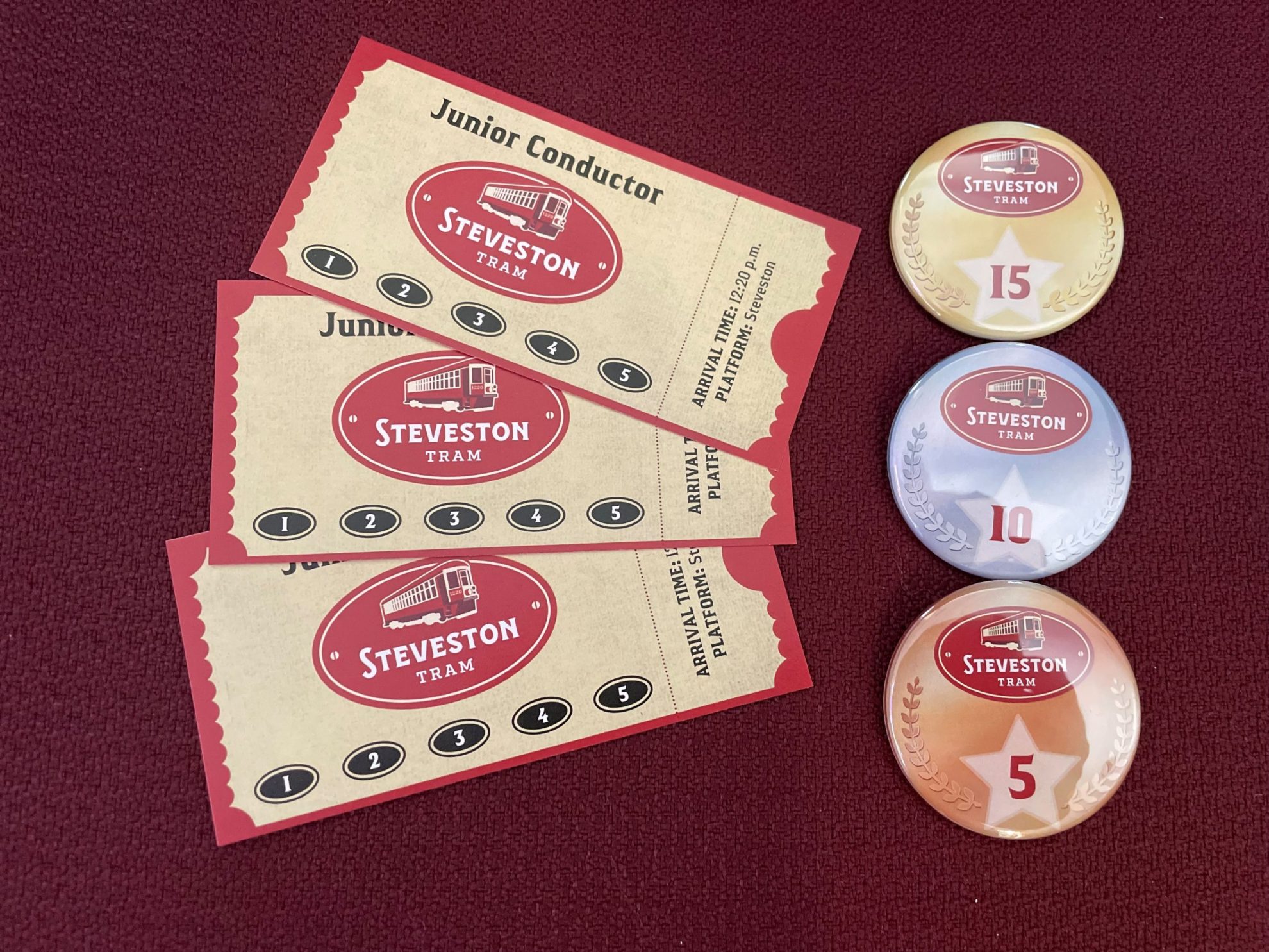 Three Steveston Tram Junior Conductor tickets and three button badges laid on a dark red background