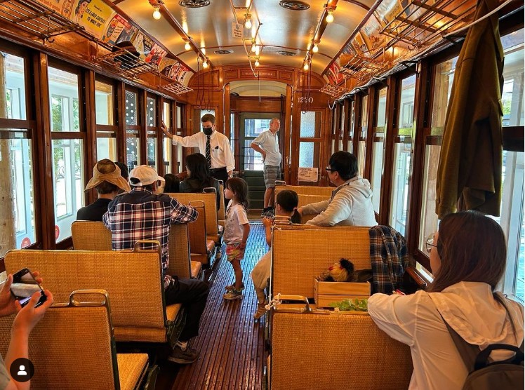 Interior of historic tram with various people seated and man with white shirt and dark tie talking to the group
