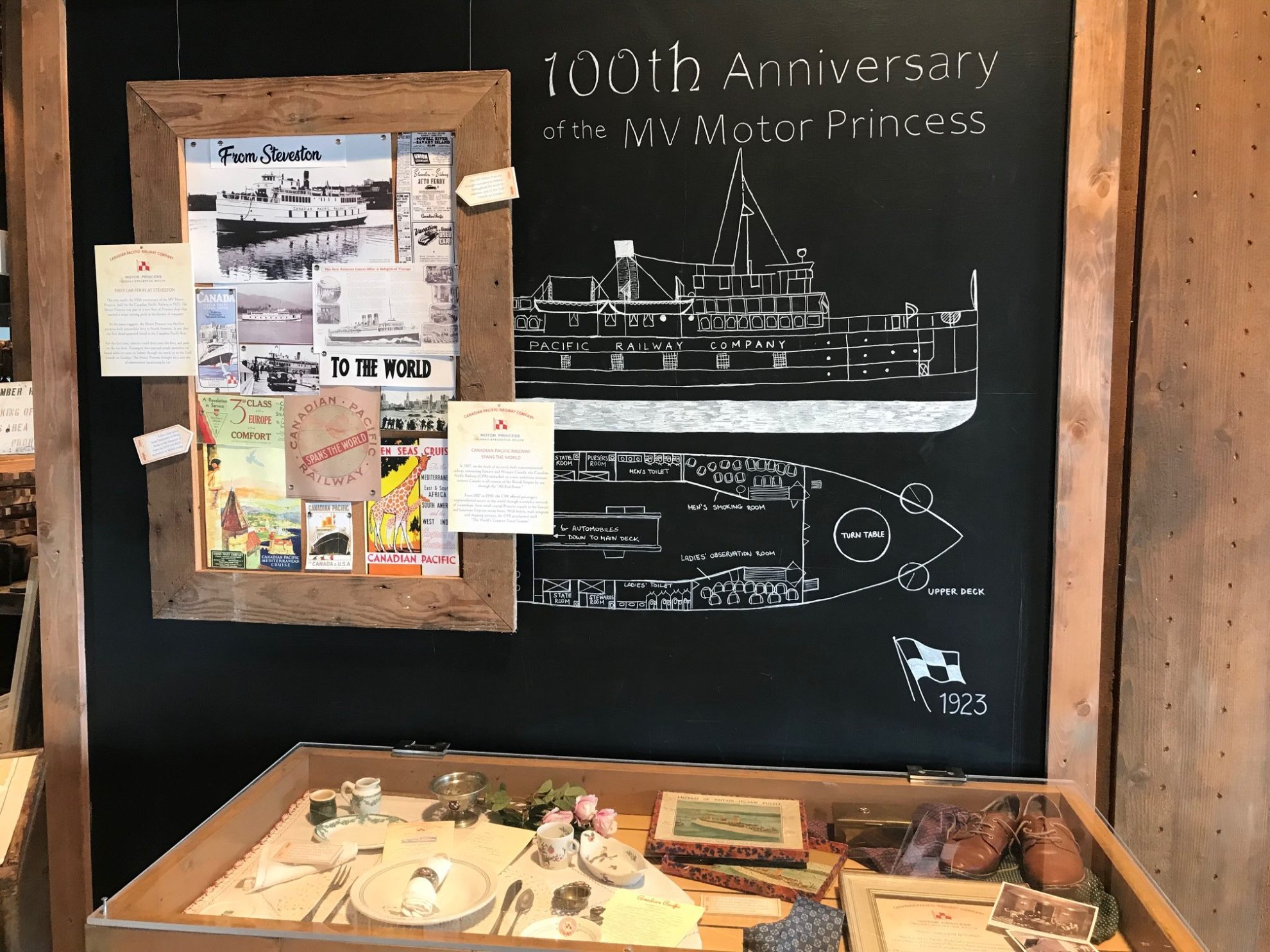 Chalkboard with side view and cross-section chalk drawing of the MV Motor Princess next to a bulletin board of archival documents and displayed above plexi covered table displaying a dining set and museum artefacts