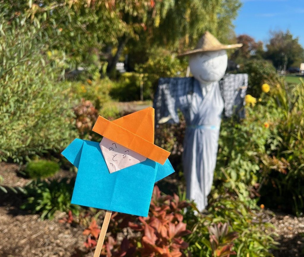 Origami paper kakashi scarecrow with blue body and orange hat held up in front of a kakashi in background