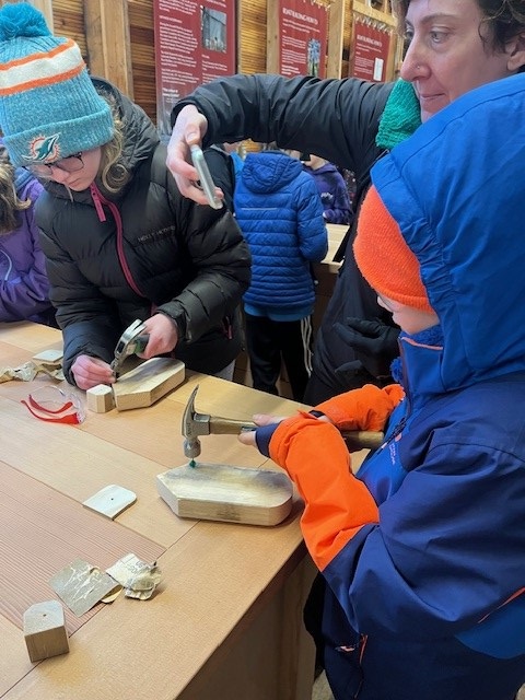 Boy in blue jacket with orange toque and gloves, building a small wooden boat