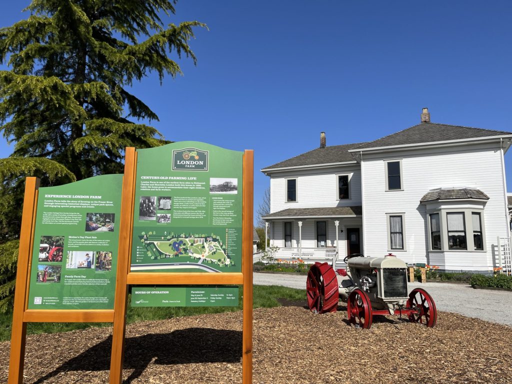Green sign in front of white tractor with red tires, and white London Farmhouse in Background