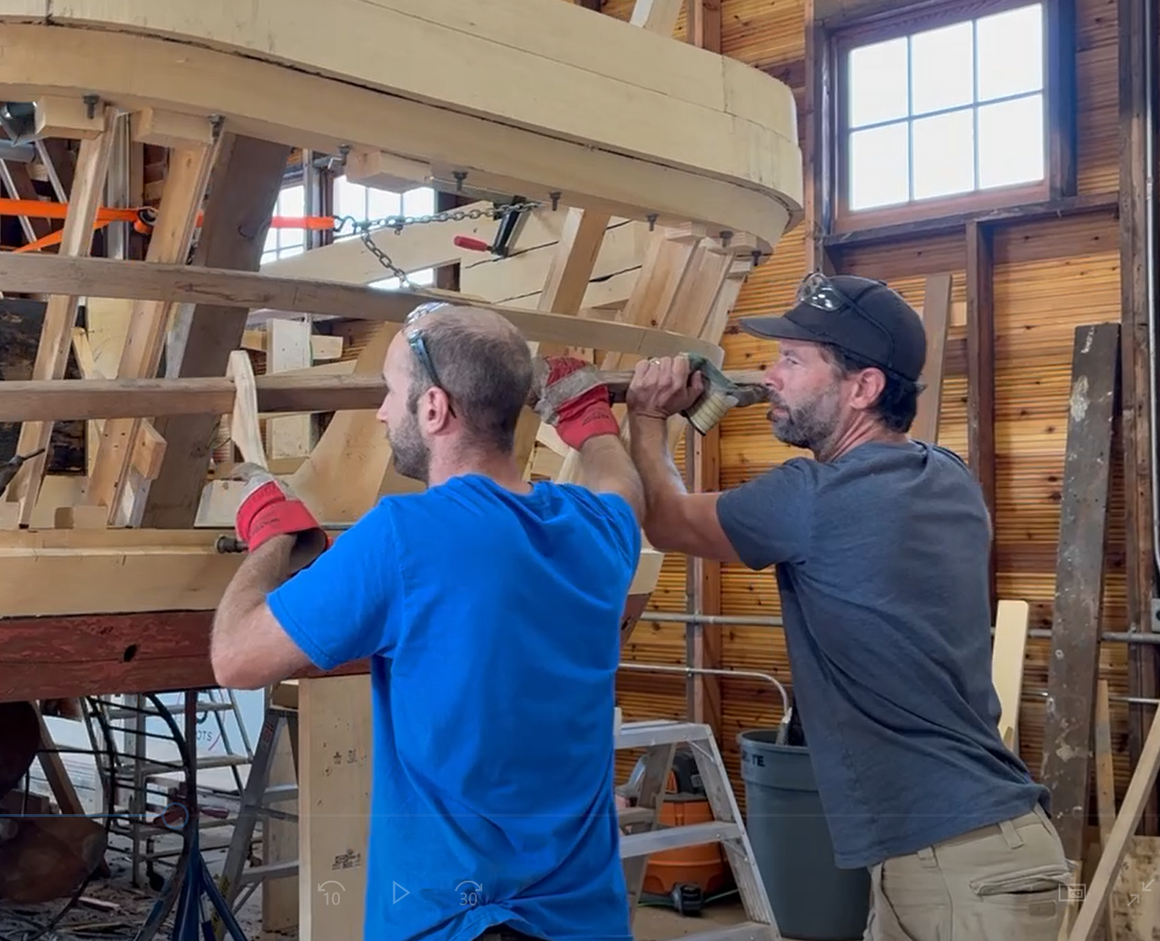Two men in blue tshirts bending wood around wooden boat ribs