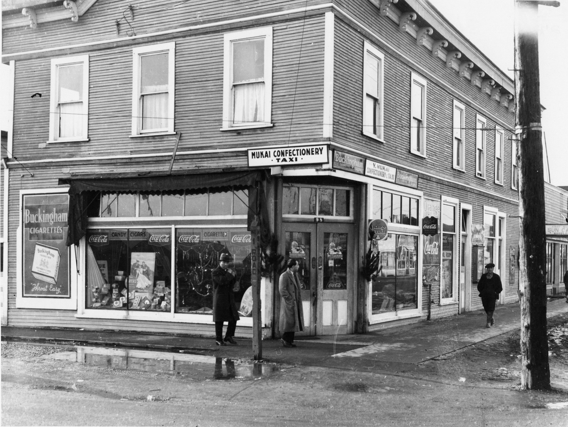 Black and white photos of men standing in front of a two storey wooden building on a street corner. Building sign says Mukai Confectionery.