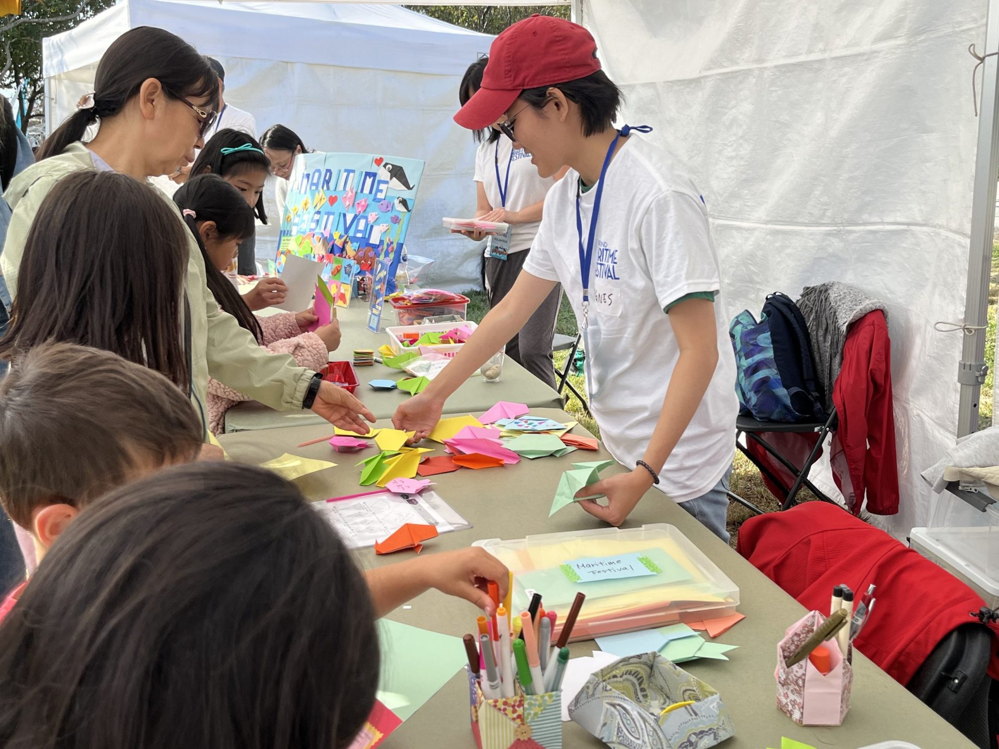 Woman in red cap and white tshirt standing at table with origami activities in front of a family