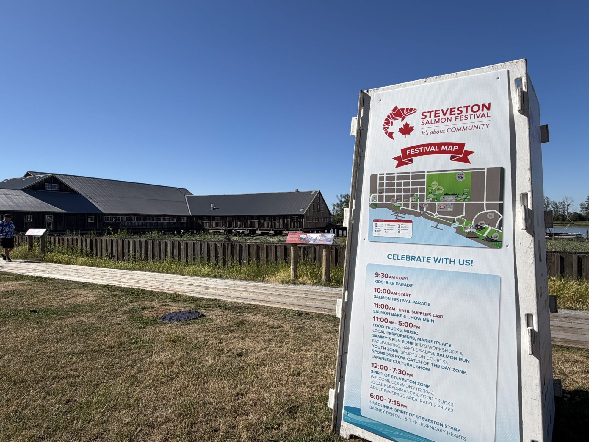 Wooden kiosk with Salmon Festival information on field with wooden shipyard building in background